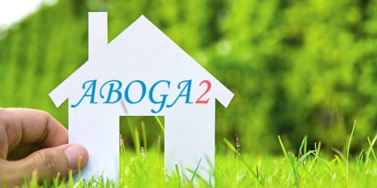Mano sosteniendo una casa de papel con el texto "ABOGA2" en un entorno verde.